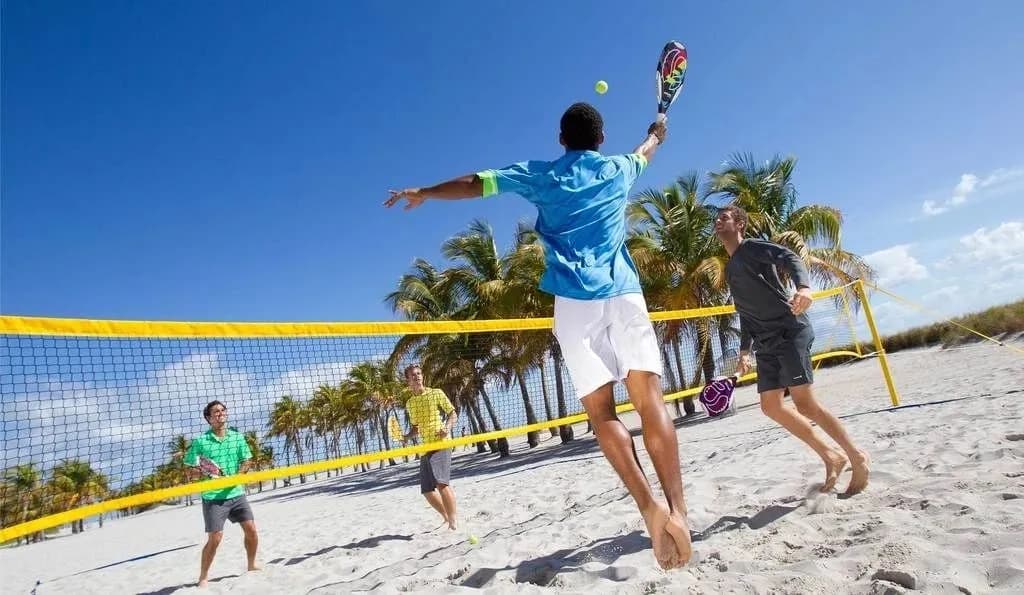 A wide shot of a beach tennis match in progress with a sunset in the background.